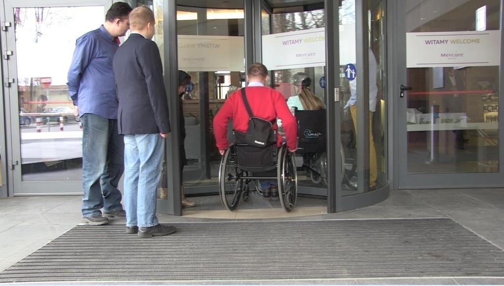 Przykład Drzwi obrotowe i umieszczone po bokach drzwi rozwierane. Drzwi obrotowe dodatkowo wyposażono w przycisk zwalniający prędkość obrotu dla osób z niepełnosprawnościami.