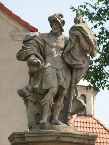 Florian ubrany w zbroję i hełm, w prawej ręce trzyma skopek, z którego wylewa się woda na płonącą budowlę, w lewej dzierży chorągiew.