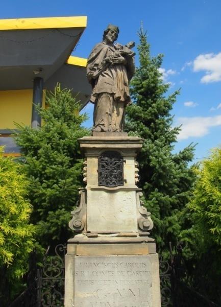 Posąg stoi na ozdobnym postumencie, ujętym w górnej części esownicami zdobionymi kampanulą z przeźroczem z dwóch stron.