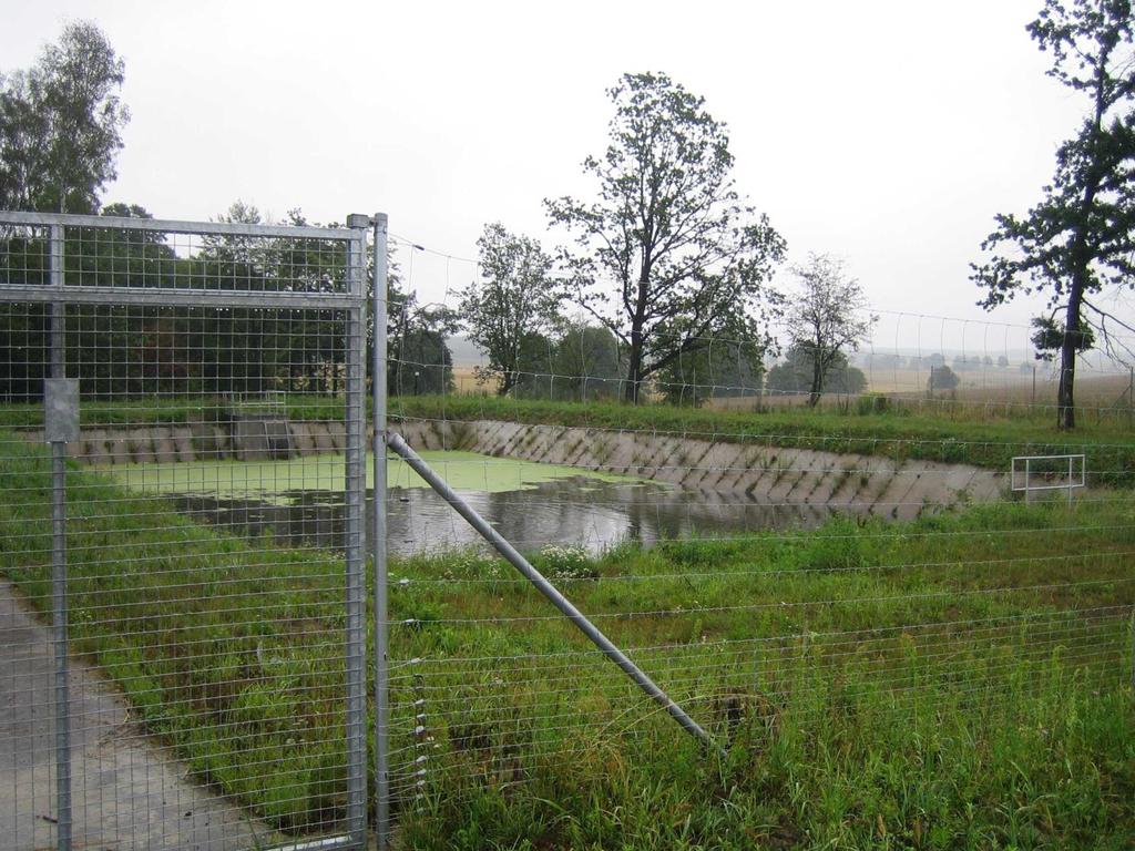 Otwarty zbiornik retencyjny ogrodzony siatką i z betonowymi skarpami na tle lasów i pól Wymagane liczne działania