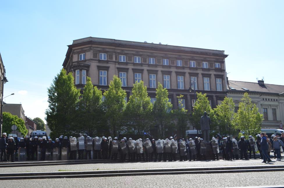 duży wóz policyjny z armatką wodną i taranem oraz 9 zwykłych wozów policyjnych. Część funkcjonariuszy trzymało pałki szturmowe w gotowości.