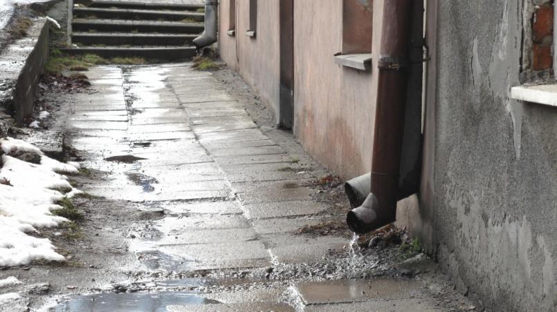 zaniedbany chodnik przy krawędzi jezdni, zalewany wodą opadową; strefy wejściowe do budynków