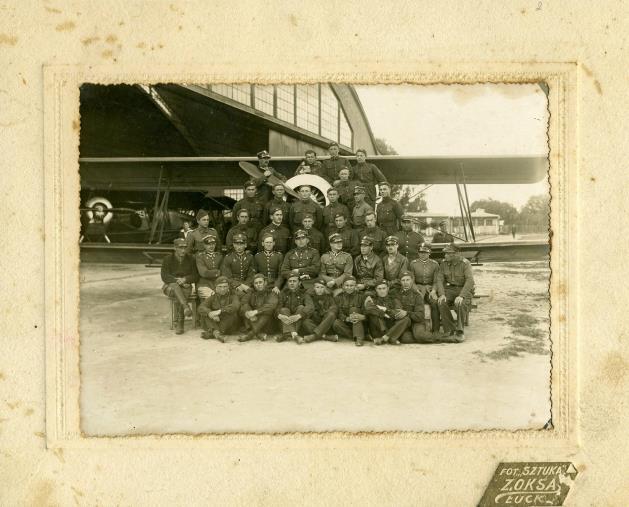 Fotografia na tekturze przedstawiająca pilotów instruktorów, podoficerów personelu naziemnego oraz kursantów paramilitarnego Przysposobienia Wojskowego Lotniczego przed hangarem na lotnisku w Łucku