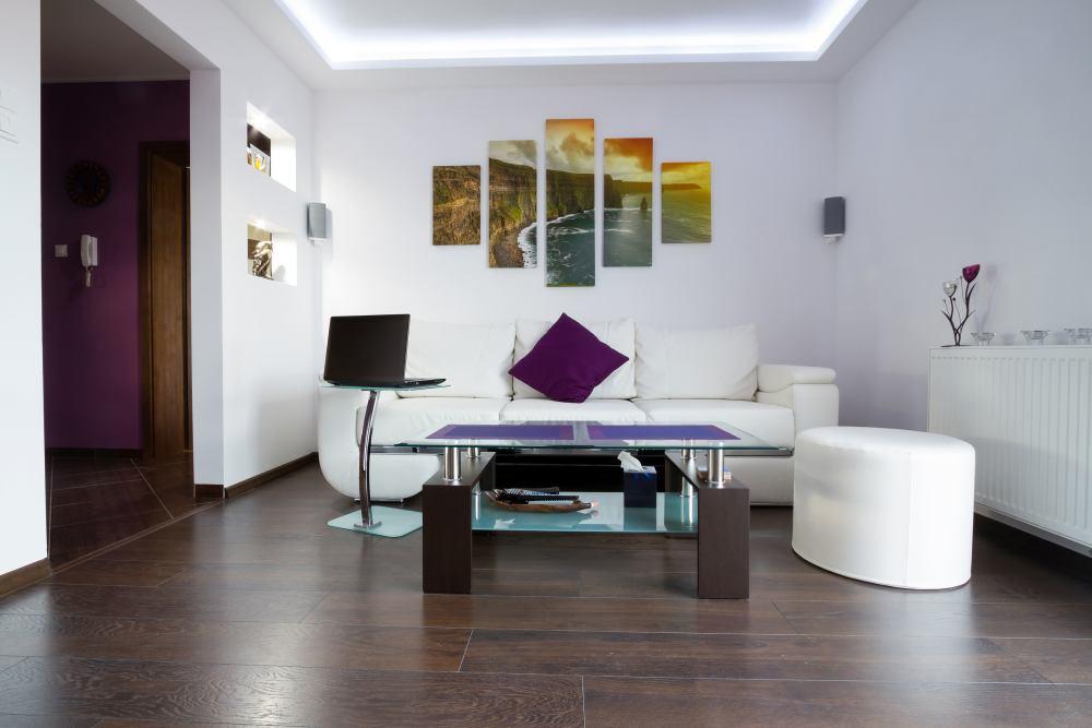 Biała, nowoczesna sofa trzyosobowa uzupełniona o pufę stanowi główny element strefy wypoczynkowej. Dzięki jasnej kolorystyce mebel - mimo pokaźnych rozmiarów - nie przytłacza wnętrza.